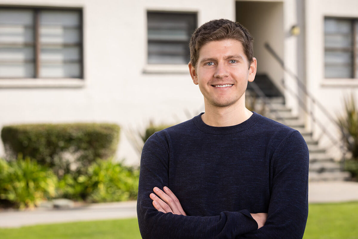 Smiling man in a navy sweater standing with his arms crossed outside a residential building.